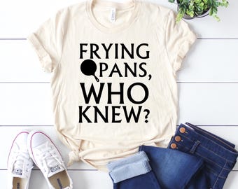 May include: A beige t-shirt with black text that reads "FRYING PANS, WHO KNEW?" with a black frying pan icon. The t-shirt is laying on a white surface with a pair of white sneakers and blue jeans.