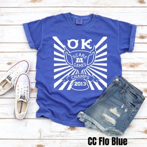 May include: A blue t-shirt with a white graphic of a trophy with the text "OK SCARE GAMES CHAMPS 2013".