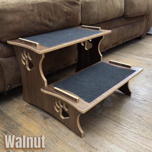 May include: A two-step pet stair made of walnut-stained wood. Each step has a dark gray, non-slip surface. Paw print cutouts decorate the sides. The word "Walnut" is printed at the bottom.