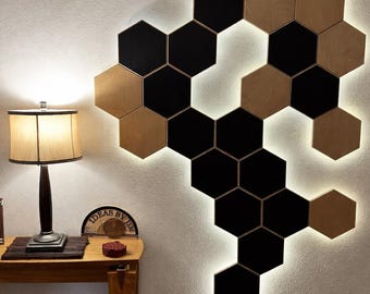 May include: A wall-mounted art installation composed of black and light wood-toned hexagonal panels. The panels are arranged in a honeycomb pattern, with a soft, glowing light outlining the design. A table lamp and side table are visible in the foreground.