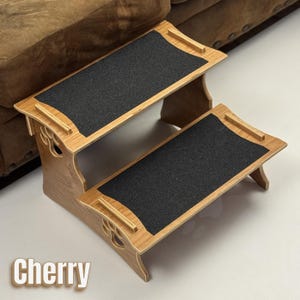 May include: Cherry-colored, two-step pet stairs with black, textured treads. The wooden frame features decorative cutouts and small handles. The stairs are designed to help pets access furniture. The word "Cherry" is displayed at the bottom.