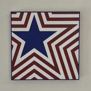 May include: Square artwork featuring a blue star at the center, surrounded by red and white stripes. The design is reminiscent of the American flag, with a textured surface and a dark blue border. The artwork is square and appears to be a decorative piece.