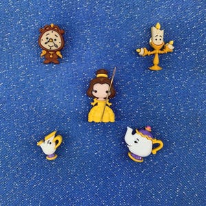 May include: A collection of colorful character-shaped embellishments from the animated film, Beauty and the Beast. The set includes a clock, a candlestick, a teapot, a teacup, and Belle in her yellow gown, all on a blue background.