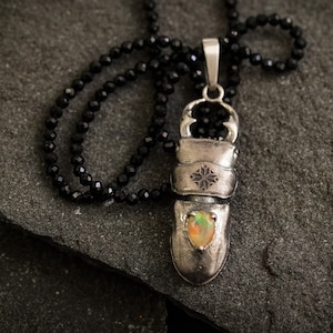 May include: A silver pendant necklace with a black spinel chain. The pendant is shaped like a beetle with a small opal stone in the center.
