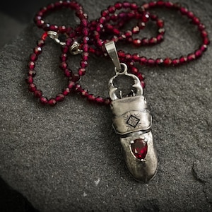 May include: A silver pendant necklace with a red gemstone. The pendant is shaped like a beetle and has intricate details. The necklace is made of red beads.