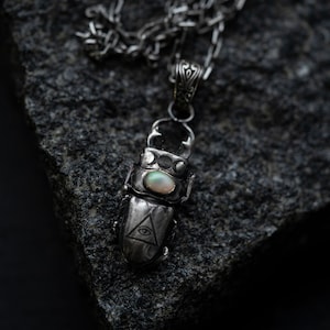 May include: A silver necklace with a beetle pendant featuring an opal stone and a triangle with an eye design. The pendant is hanging on a dark gray rock.