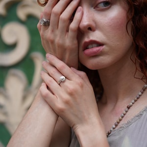 May include: A silver ring with a round, clear stone is worn on a finger. Another ring with a textured design is also visible. A beaded necklace and a light-colored top are also in the image.