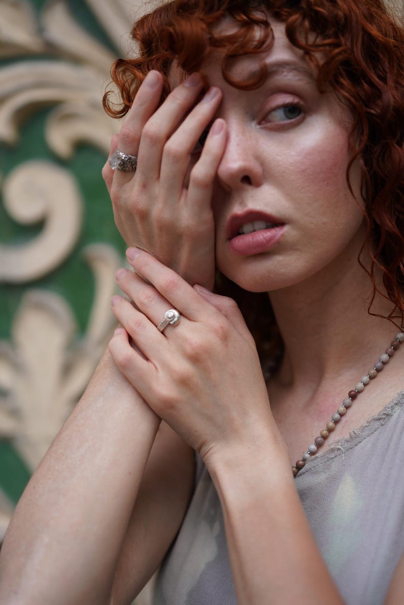 May include: A silver ring with a round, clear stone is worn on a finger. Another ring with a textured design is also visible. A beaded necklace and a light-colored top are also in the image.