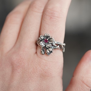 Könnte beinhalten: Ein silberner Ring mit floralem Design und einem zentralen roten Edelstein. Der Ring hat detaillierte Blütenblätter und Knospen, die eine von der Natur inspirierte Ästhetik schaffen. Der Ring wird an einem Finger getragen.