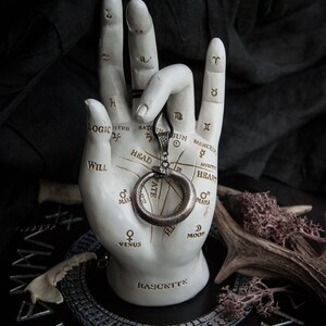 May include: A silver snake ring pendant with a circular design, hanging from a black cord. The pendant is displayed on a white hand with a palmistry chart, resting on a black circular base with silver runes.