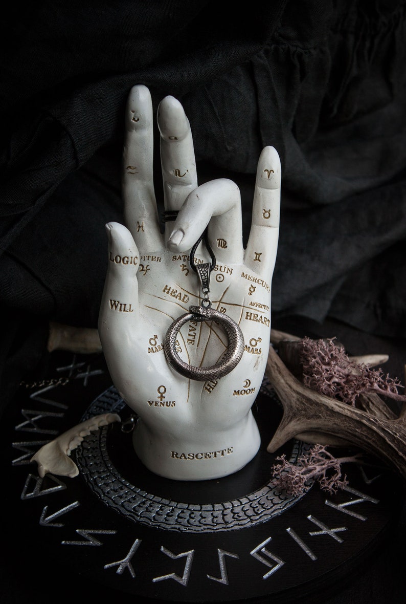 May include: A silver snake ring pendant with a circular design, hanging from a black cord. The pendant is displayed on a white hand with a palmistry chart, resting on a black circular base with silver runes.