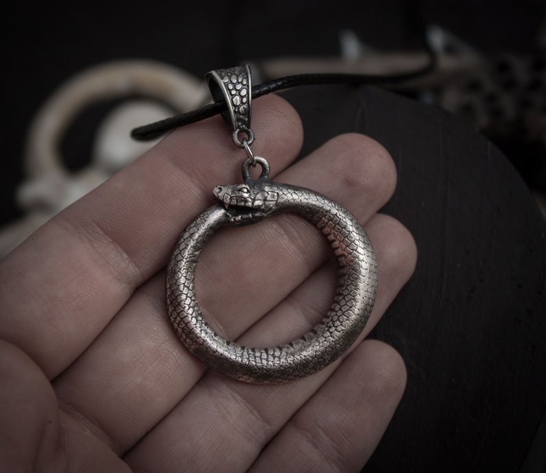 May include: Silver pendant necklace featuring a snake forming a circle with its tail in its mouth. The snake is detailed with scales and has a realistic head.