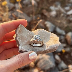 May include: A silver ring with a textured band and a spiral design on a stone. The ring features a small, clear gemstone at the center of the spiral. The ring is held in a hand with light pink nail polish.