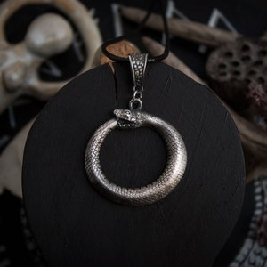 May include: A silver snake pendant necklace with a snake forming a circle. The pendant is on a black surface.