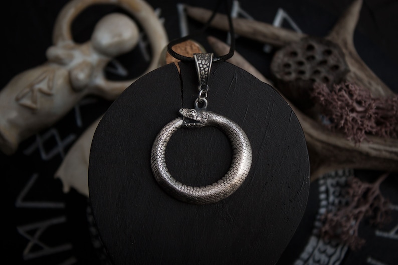 May include: A silver snake pendant necklace with a snake forming a circle. The pendant is on a black surface.