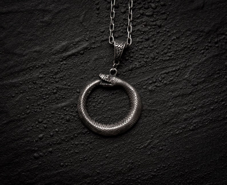 May include: A silver necklace with a snake pendant in the shape of a circle. The snake is biting its own tail, forming a continuous loop.