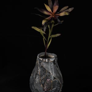 Puede incluir: Un jarrón de cerámica gris oscuro con una superficie texturizada, que sostiene una flor con pétalos morados oscuros y amarillos. El jarrón tiene detalles dorados y un elemento similar a un cristal púrpura. El fondo es negro.