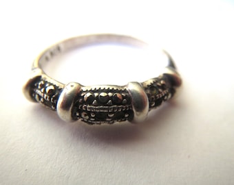 May include: A silver ring featuring a band with three distinct sections separated by smooth metal bars. Each section is inlaid with small, dark, faceted stones that create a textured, metallic appearance across the front of the ring.