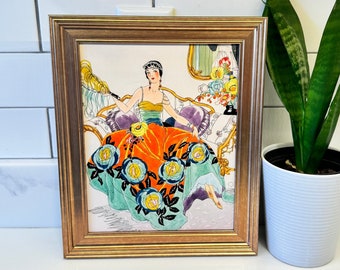 May include: A framed illustration of a woman in a colorful dress with floral accents. The woman is sitting on a couch with a decorative throw pillow. The illustration is set against a light background.