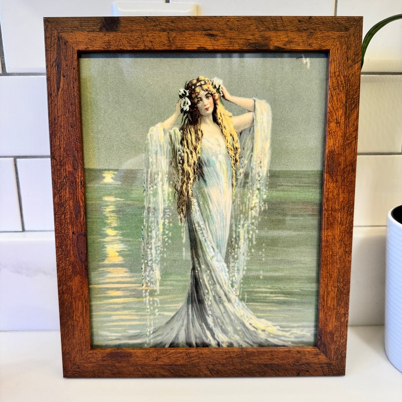 K&ouml;nnte beinhalten: Eine gerahmte Vintage-Illustration einer Frau in einem flie&szlig;enden wei&szlig;en Kleid, die im Meer steht. Sie hat langes blondes Haar und tr&auml;gt eine Krone aus wei&szlig;en Blumen. Das Wasser glitzert und der Himmel ist zartblau.