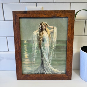 K&ouml;nnte beinhalten: Ein gerahmter Vintage-Druck einer Frau in einem flie&szlig;enden wei&szlig;en Kleid, die im Meer steht. Die Frau hat langes blondes Haar und tr&auml;gt eine Blumenkrone. Das Meer ist hellblaugr&uuml;n und der Himmel ist hellblau.