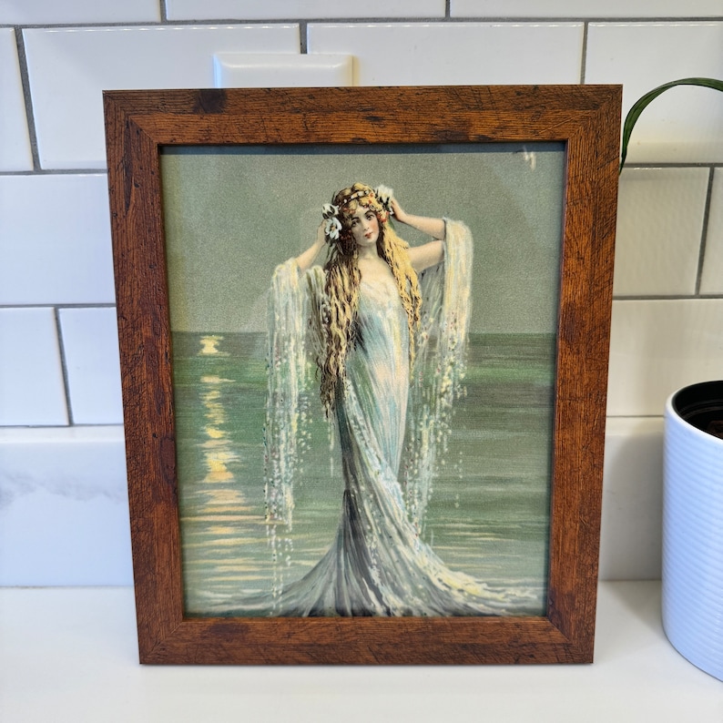 K&ouml;nnte beinhalten: Ein gerahmter Vintage-Druck einer Frau in einem flie&szlig;enden wei&szlig;en Kleid, die im Meer steht. Die Frau hat langes blondes Haar und tr&auml;gt eine Blumenkrone. Das Meer ist hellblaugr&uuml;n und der Himmel ist hellblau.