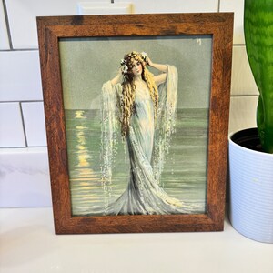 K&ouml;nnte beinhalten: Eine gerahmte Vintage-Illustration einer Frau mit langen blonden Haaren, die ein wei&szlig;es Kleid und eine Blumenkrone tr&auml;gt und im Meer steht, mit Wellen und Mondlicht im Hintergrund.