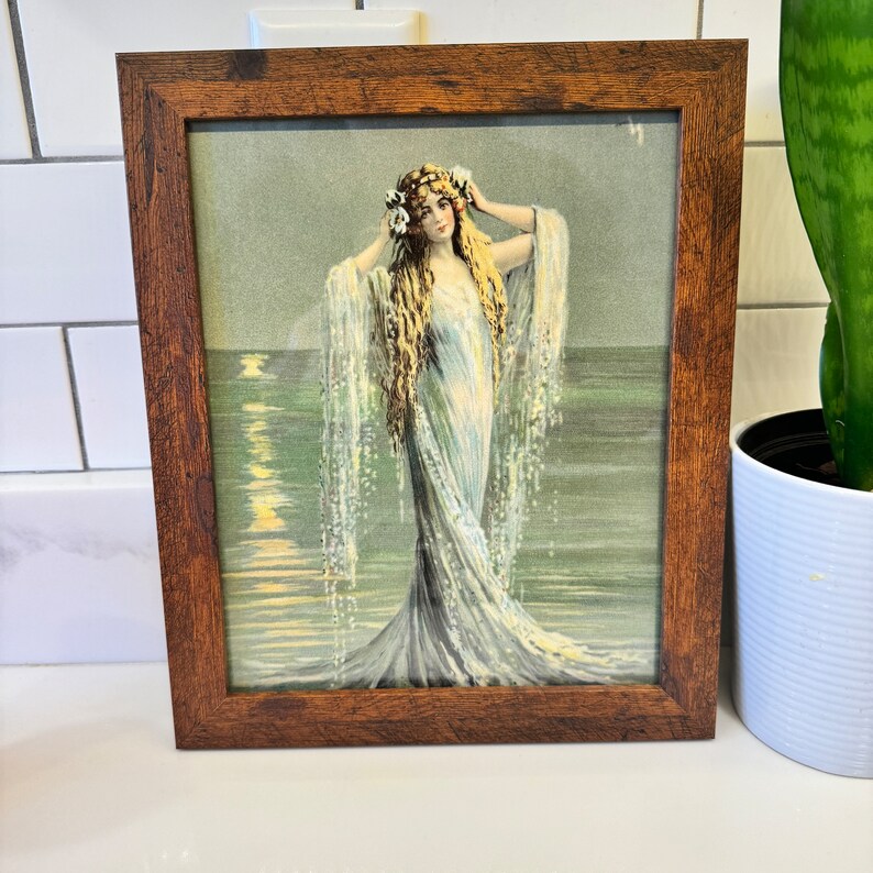 K&ouml;nnte beinhalten: Eine gerahmte Vintage-Illustration einer Frau mit langen blonden Haaren, die ein wei&szlig;es Kleid und eine Blumenkrone tr&auml;gt und im Meer steht, mit Wellen und Mondlicht im Hintergrund.