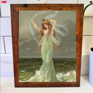 May include: A framed vintage print of a woman in a flowing green dress standing in the ocean. The woman has long brown hair and is holding a sheer white scarf. The frame is made of dark brown wood.