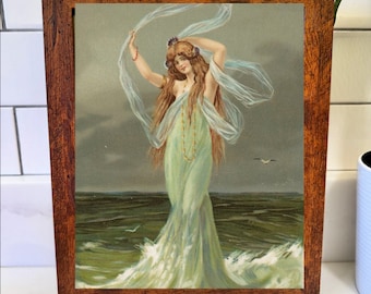May include: A framed vintage print of a woman in a flowing green dress standing in the ocean. The woman has long brown hair and is holding a sheer white scarf. The frame is made of dark brown wood.