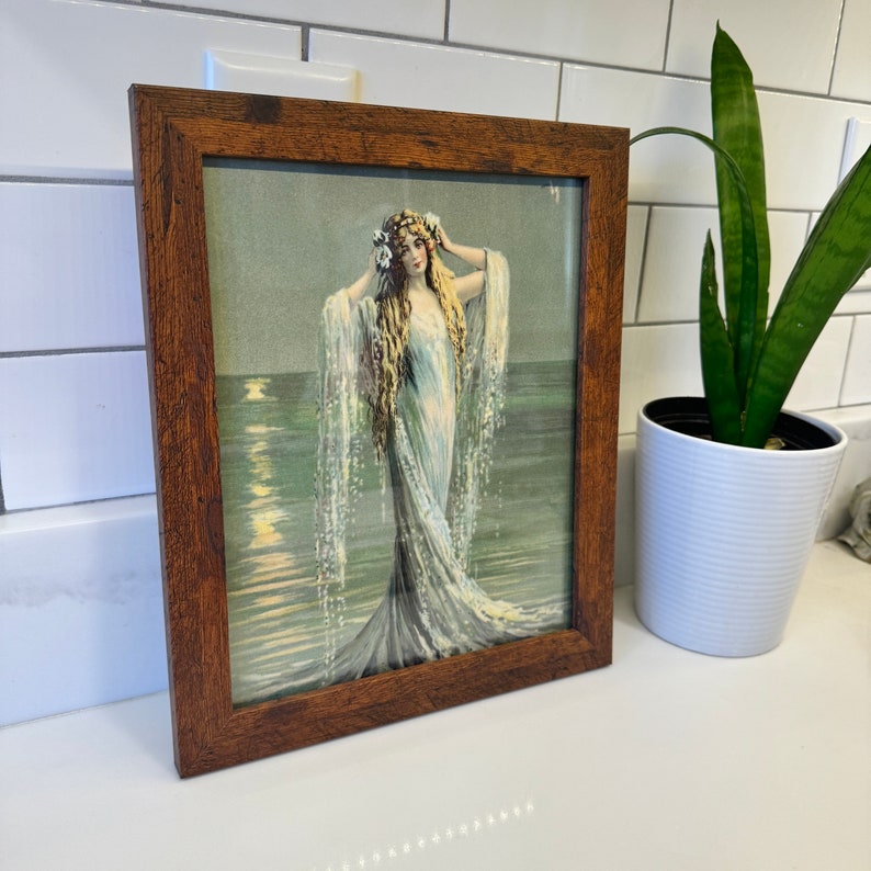 K&ouml;nnte beinhalten: Eine gerahmte Vintage-Illustration einer Frau in einem flie&szlig;enden wei&szlig;en Kleid, die im Meer steht, mit einem mondbeschienenen Himmel im Hintergrund.