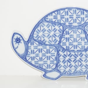 May include: A ceramic turtle-shaped dish with a blue and white geometric pattern. The dish has a detailed design on the shell and a smiling face on the head. The dish is a decorative item.