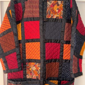May include: A long-sleeved quilted jacket featuring a patchwork design of square fabric panels in shades of red, orange, brown, and dark blue. The jacket has a round neckline, black fabric trim between the panels, and three dark toggle closures.