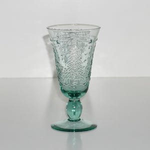 May include: A single, clear teal-colored glass with a textured pattern. The glass has a stem and a round base. The glass is likely used for beverages and is set against a white background.