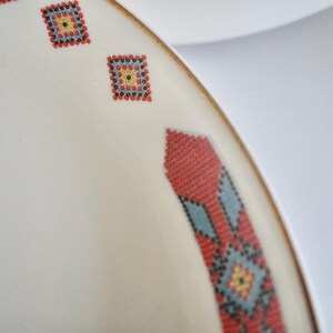 May include: Close-up of a white ceramic plate with a decorative border. The border features a repeating pattern of small, colorful, square designs in red, blue, and yellow. The plate has a gold rim.