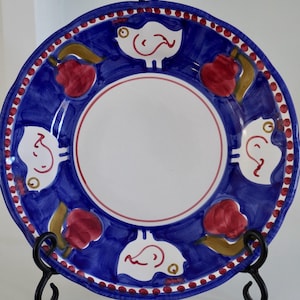May include: A decorative ceramic plate with a blue border featuring white bird and red fruit designs. The plate has a white center and a red dotted border. The plate is displayed on a black metal stand.