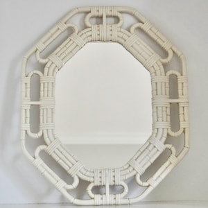 May include: An octagonal wall mirror featuring a cream-colored frame constructed from woven rattan. The frame displays a geometric pattern with interconnected loops and straight segments, surrounding a central reflective glass surface.