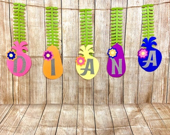 May include: A colorful banner spelling "DIANA" with pineapple-shaped cutouts. Each cutout is a different color: pink, orange, yellow, purple, and blue. Each pineapple has a silver letter and a flower accent. The banner is suspended by a string.