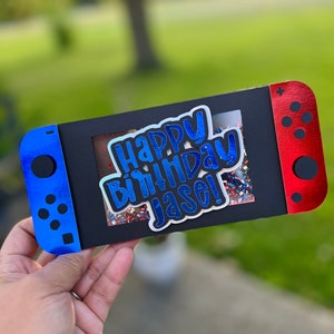 May include: A black card shaped like a game console controller with blue and red accents. The card has the text "Happy Birthday Jase!" in blue and white, with a confetti-filled window in the center. The controller-shaped card is held in a hand.