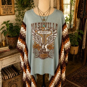 Puede incluir: Una camiseta azul claro con un gráfico de una guitarra con alas y el texto Nashville Music City Rock Star Tennessee. La prenda tiene paneles laterales de ganchillo en marrón, beige y blanco, exhibida en un maniquí con un collar de capas.