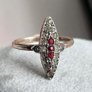 May include: A vintage ring with a silver band and a central, elongated design. The design features a row of three red gemstones flanked by smaller, clear stones. The ring is set on a gray, textured surface.