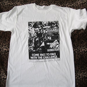 Könnte beinhalten: Weißes T-Shirt mit einer schwarz-weißen Grafik einer Person in Militäruniform. Die Grafik enthält Text: "CHAIN X OF STRENGTH", "CARRY NATION", "INSIDE OUT", "FORCED DOWN" und "COME FACE TO FACE WITH THE CHALLENGE."