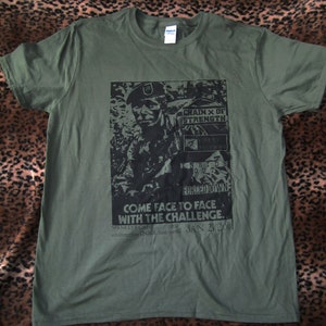 Puede incluir: Una camiseta verde con un estampado gráfico en blanco y negro de un soldado. El gráfico incluye el texto "Chain X of Strength", "Inside Out", "Forced Down", y "Come Face to Face with the Challenge". El texto "Spankeys Cafe" y "Jan 21 2001" también están incluidos en el gráfico.