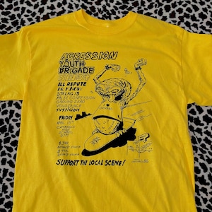 May include: Yellow t-shirt with a black graphic print of a cartoon alien skateboarding. The graphic includes the text "Agresson Youth Brigade Sin3+", "Ill Repute Dr Know", "Stalag 13", "False Confession", "Ground Zero", "Vengeance", "Suspicion", "Oxnard", "Friday April 15th", "Camarillo High School Gym", "$3.00 before 7 o'clock", "$4.00 after 7 o'clock", and "Support the Local Scene!"