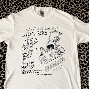 May include: White t-shirt with a black and white graphic print of a skeleton skateboarding. The graphic includes the text "L.A.s First All Skate Gig!" with "The Big Boys", "J.F.A. Aggression Faction", "Ill Repute", "Tales of Terror", "Friday 7:30 May 18th", "Stardust Ballroom 5612 Sunset Blvd", and "For Info 464-2205".