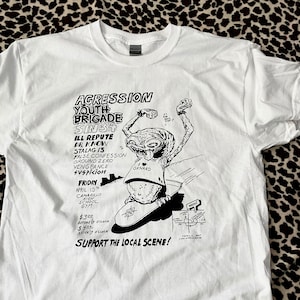 May include: White t-shirt with a black and white illustration of a cartoon alien skateboarding. The text on the shirt reads "Agresssion Youth Brigade Sin3+" and lists a series of bands with the date and location of a show. The text also includes the price of admission: $3.00 before 7:00 p.m. and $4.00 after 7:00 p.m.