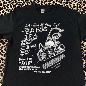 May include: Black and white t-shirt with a graphic of a skeleton skateboarding. The text on the shirt reads "L.A.s First All Skate Gig!" with "The Big Boys", "J.F.A", "Agression Faction", "Ill Repute", "Tales + Tetfor", "Friday 7:30 May 18th", "Stardust Ballroom 5612 Sunset Blvd", "For Info 464-2205", "$6.50 $8.00", "No Age Limit", and "Door Prizes PS Tracker Skates Beans and Much Much More Sponsored By Transworld Sims-Tracker-Powell Peralta-Variplex-Rip City Skates Madrid Thrasher Pro & Amatuer Fore Style Skateboarders".