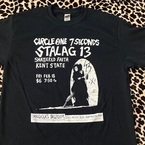 May include: Black t-shirt featuring white text and a graphic of a figure in a doorway. The text includes "CIRCLE ONE 7 SECONDS STALAG 13" and event details: "FRI FEB 18 $6 7:30 PM" and "MANDIOLA'S BALLROOM".