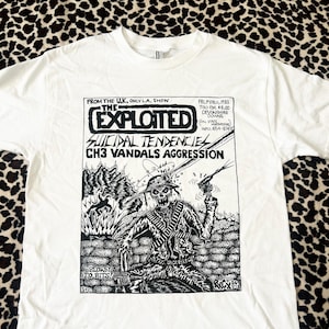 Pode incluir: Camiseta branca com uma estampa gráfica em preto e branco de um cartaz de banda. O cartaz apresenta o texto "The Exploited", "Suicidal Tendencies", "CH3 Vandals Aggression" e "Blown to Bits!" juntamente com uma ilustração em preto e branco de um esqueleto segurando uma arma.