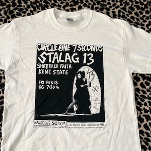 May include: White t-shirt with a black and white graphic print of a concert poster for Circle One 7 Seconds playing at the Mandirola's Ballroom in Huntington Park, California. The poster says "Stalag 13" and "Shattered Faith" with the date "Fri Feb 18" and the time "7:30 pm".
